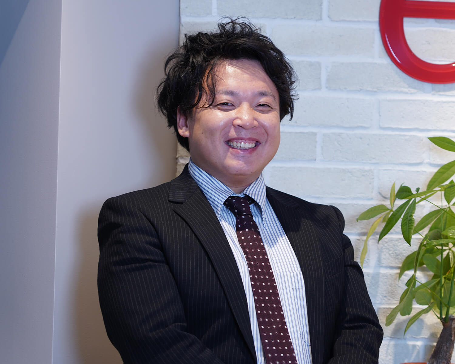 白レンガ壁を背景に、ネイビーのストライプ柄スーツ、ストライプシャツ、柄タイを着用して、手を前で重ねて微笑む日本人男性のエグゼクティブポートレート写真。