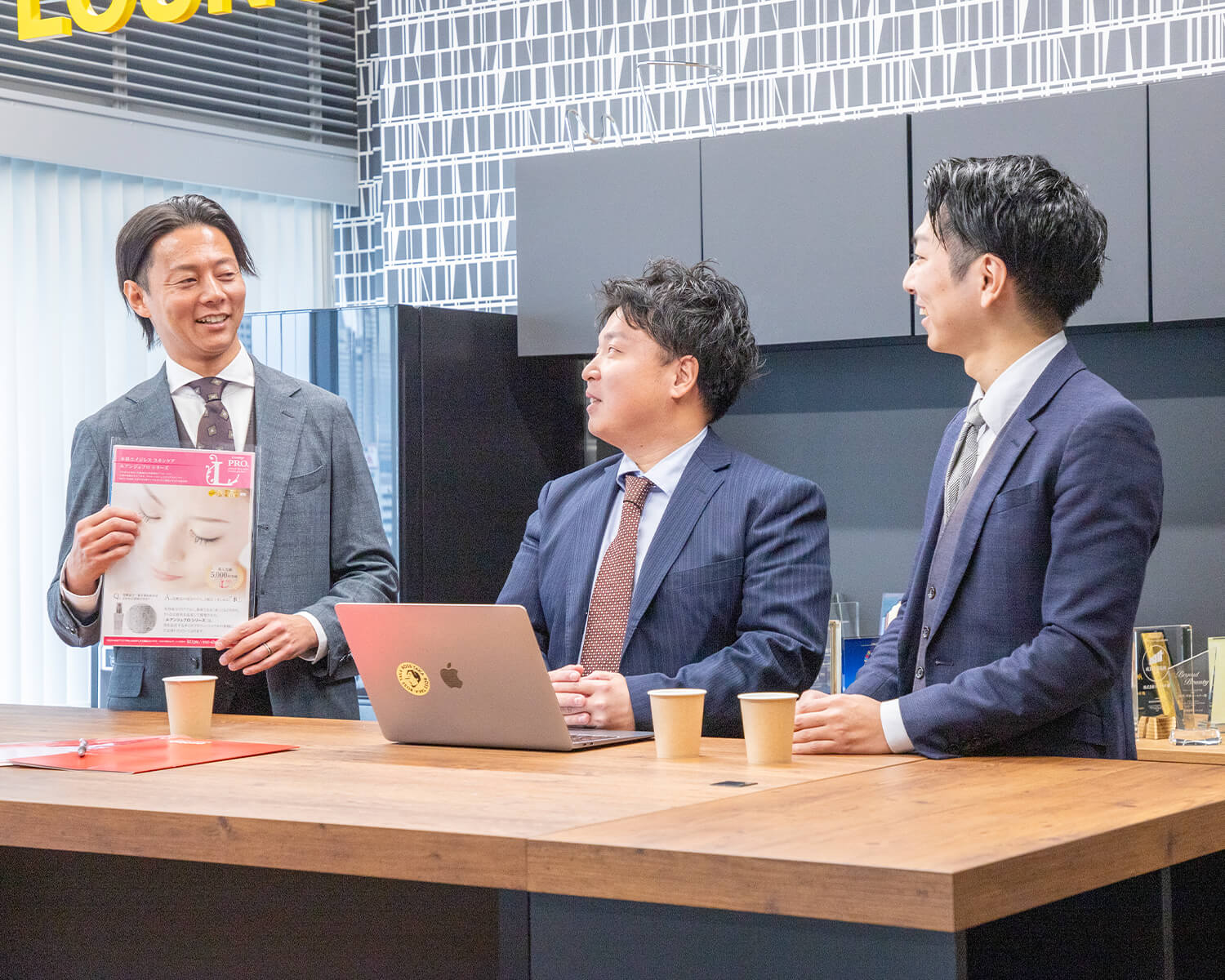 木のテーブルを囲んで会議をする3人の日本人ビジネスマン。左の男性はチラシを持ち、中央の男性はラップトップに向かい、右の男性は話を聞いている。背景はモダンなオフィス内装。