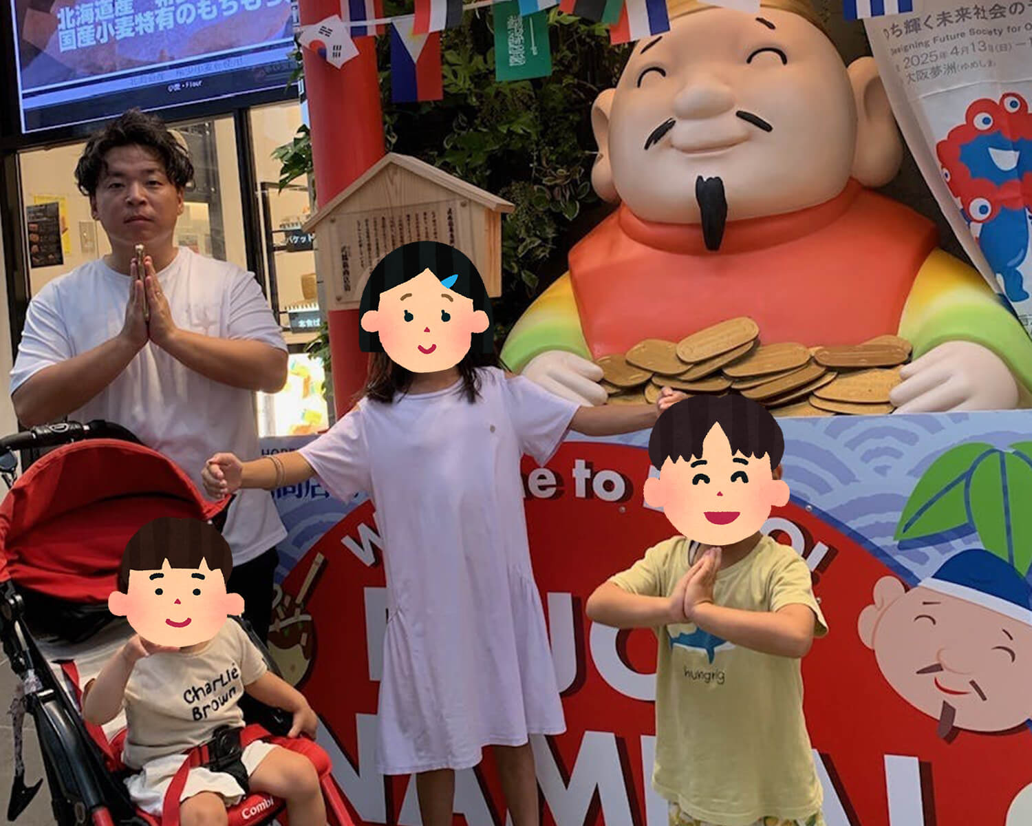 観光スポットの縁起物「えびす像」と子供たちと一緒に、白いTシャツ姿で合掌のポーズをして写る日本人男性。像の前には小判や万国旗、日本語の看板が見える。