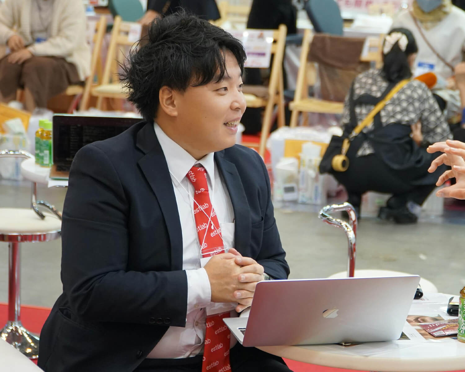 イベント会場で、黒いスーツに「estlab」とロゴの入った赤いネクタイを着用し、ラップトップが置かれた白いテーブルで、組んだ手を前に置いて微笑む日本人男性。