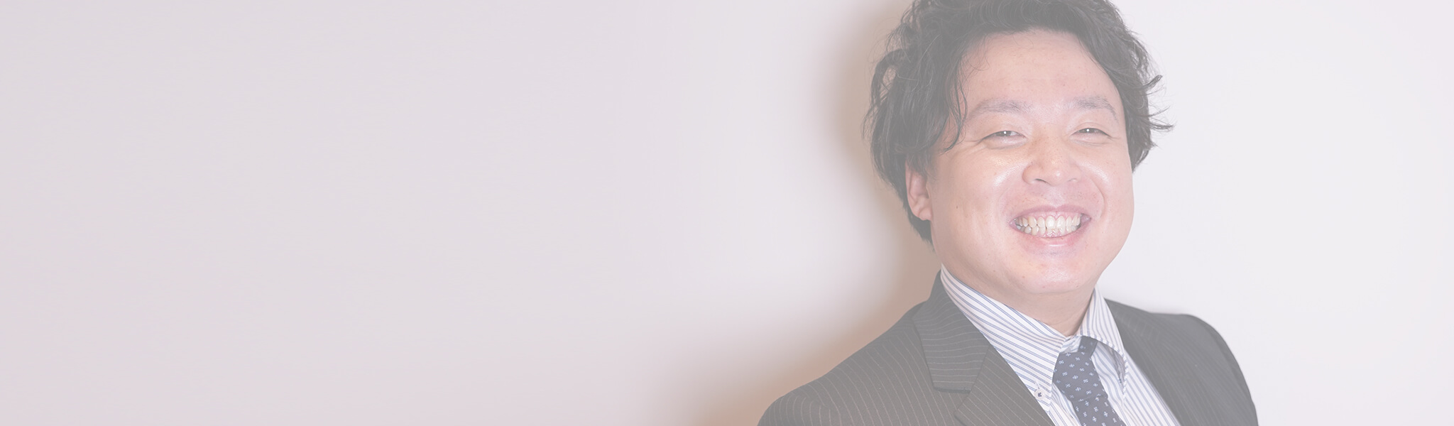 明るい光の中で微笑む日本人男性のクローズアップ。ネイビーのストライプ柄スーツ、ストライプシャツ、ドット柄のネクタイを着用している。背景はぼかしのかかった白。