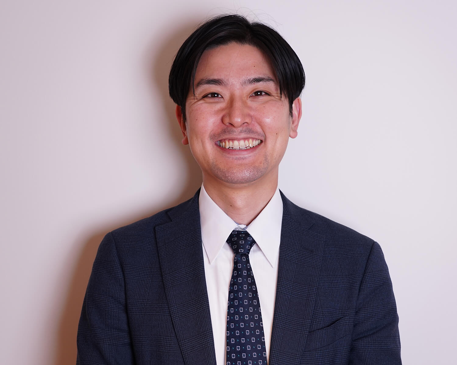 紺色のチェック柄スーツと小紋柄のネクタイを着用し、白い背景の前で爽やかに微笑むビジネスマンのバストアップ写真