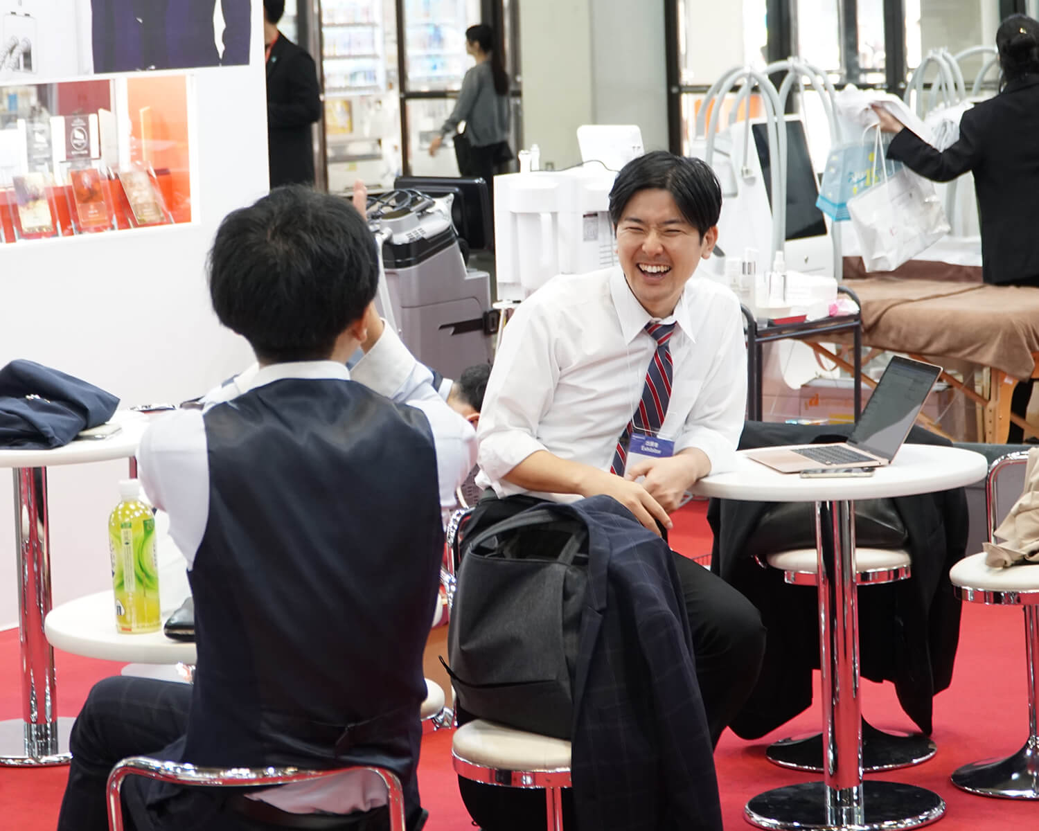 イベント会場の丸テーブルを囲み、リラックスした表情で顧客と談笑する白シャツ姿の男性。テーブルの上にはノートパソコンが置かれている
