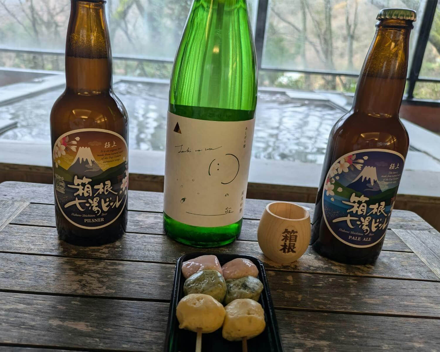 「箱根七湯ビール」のボトル2本と、可愛らしいイラストが描かれた日本酒の瓶、三色団子が並ぶ、温泉宿のテラスでのひととき