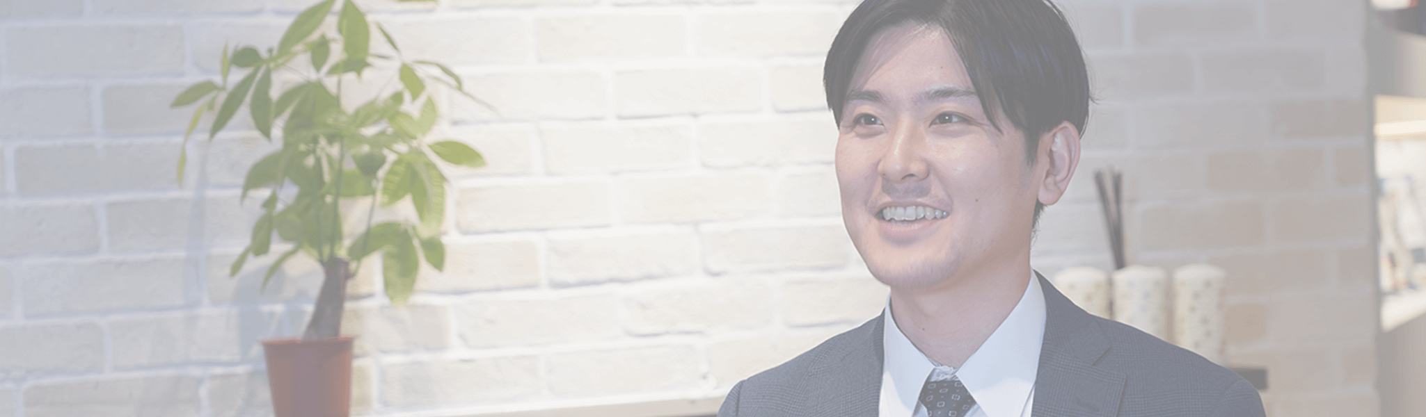 白いレンガ壁と観葉植物を背景に、明るい未来を語るような表情を見せる男性。記事のヘッダー用に淡く加工された画像