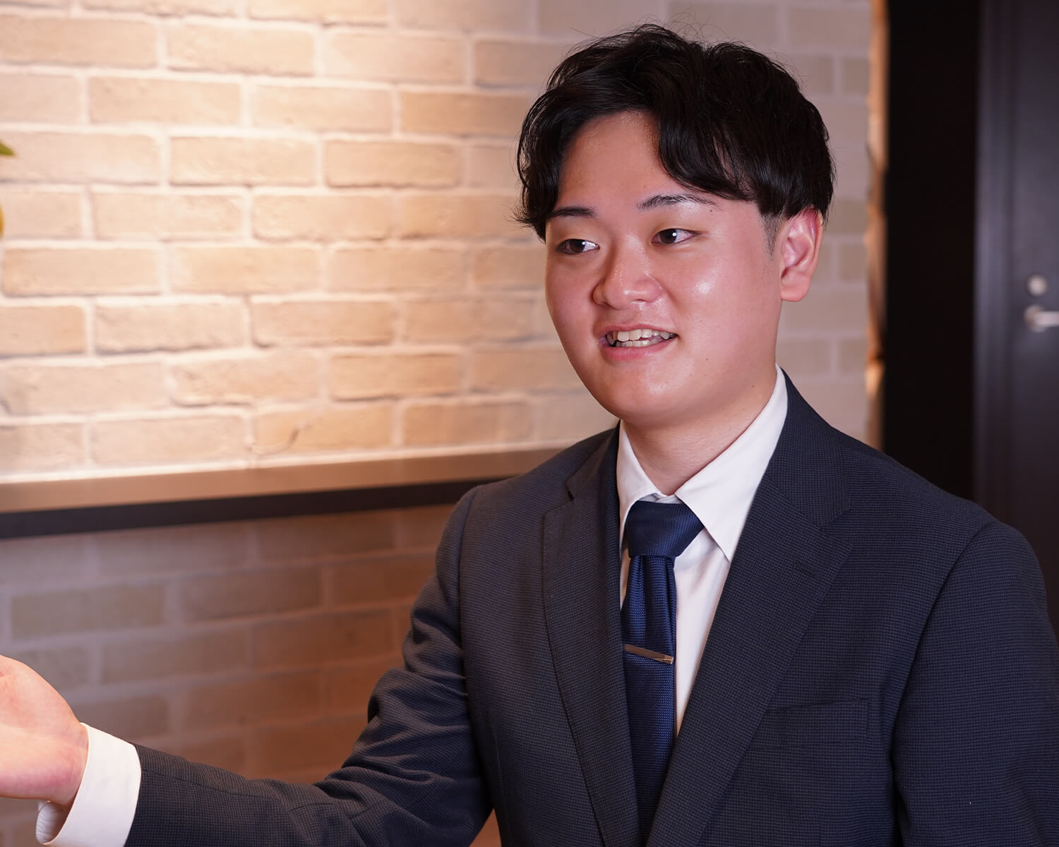 レンガ調の壁があるオフィスで、身振り手振りを交えながら話す紺色スーツの日本人男性