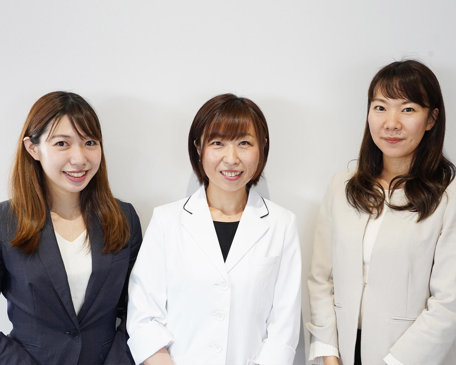 オフィスの白い壁を背に並んで立つ3人の日本人女性のポートレート。左側の女性はネイビーのジャケット、中央の女性は白衣、右側の女性はベージュのジャケットを着用しています。