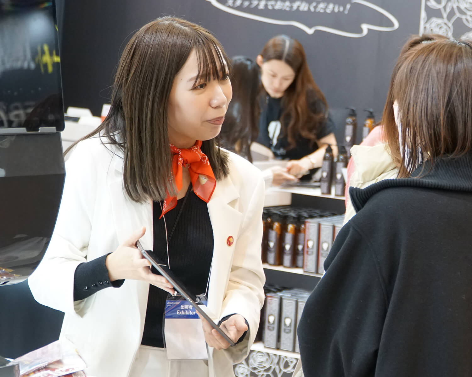 展示会のブースで、タブレットを手に来場者へ熱心に説明する白いジャケットの女性スタッフ。首元には赤いスカーフ、胸元には「Exhibitor」のバッジを着用しています。背景にはコスメ商品が陳列されています。