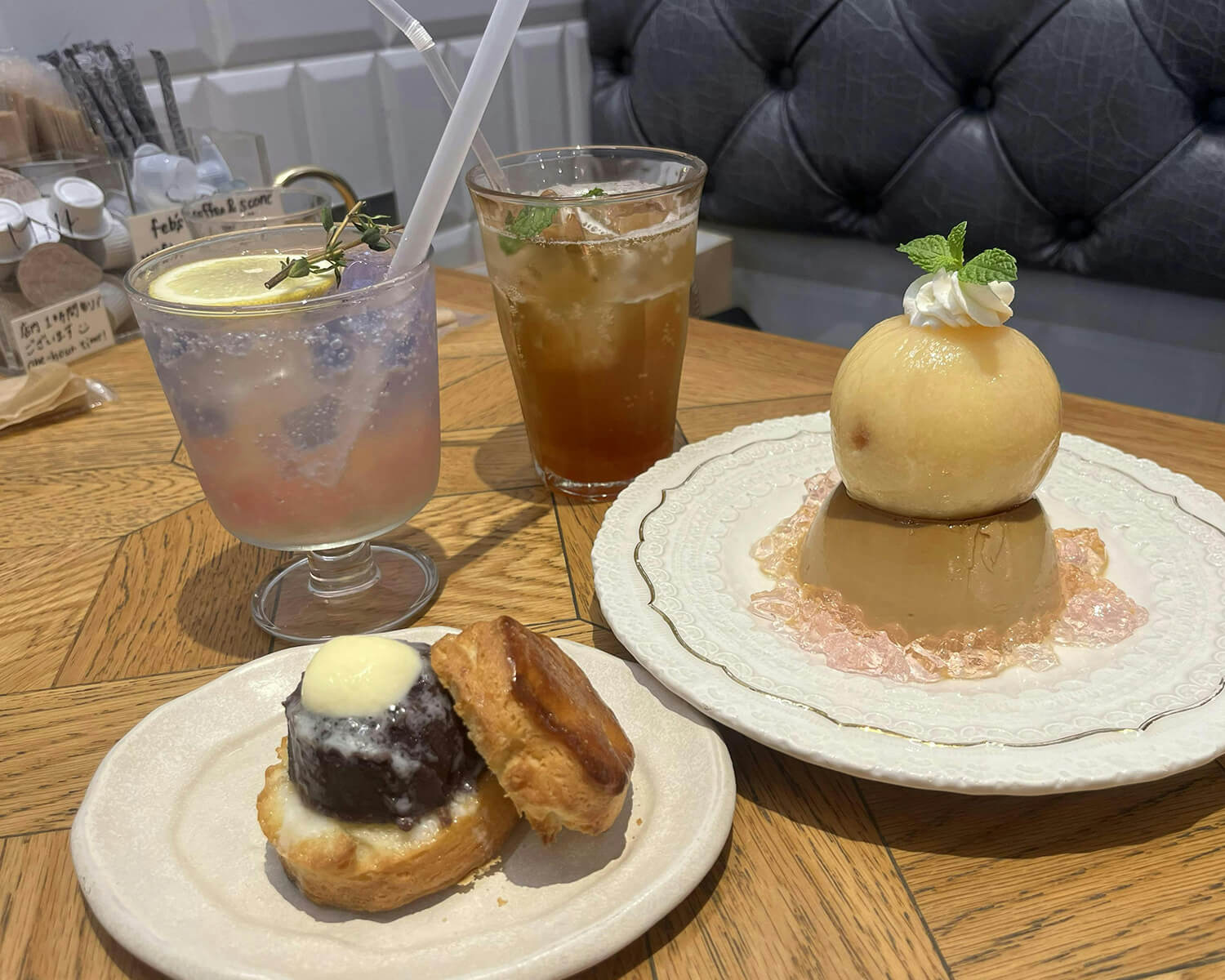 カフェの木目調テーブルに並べられた、夏らしいスイーツとドリンク。手前左には小倉バターをサンドしたスコーン、手前右には白桃が丸ごと乗ったキャラメルプリン、奥には2種類の冷たいドリンク（レモン入りバタフライピーティーとアイスティー）が置かれています。