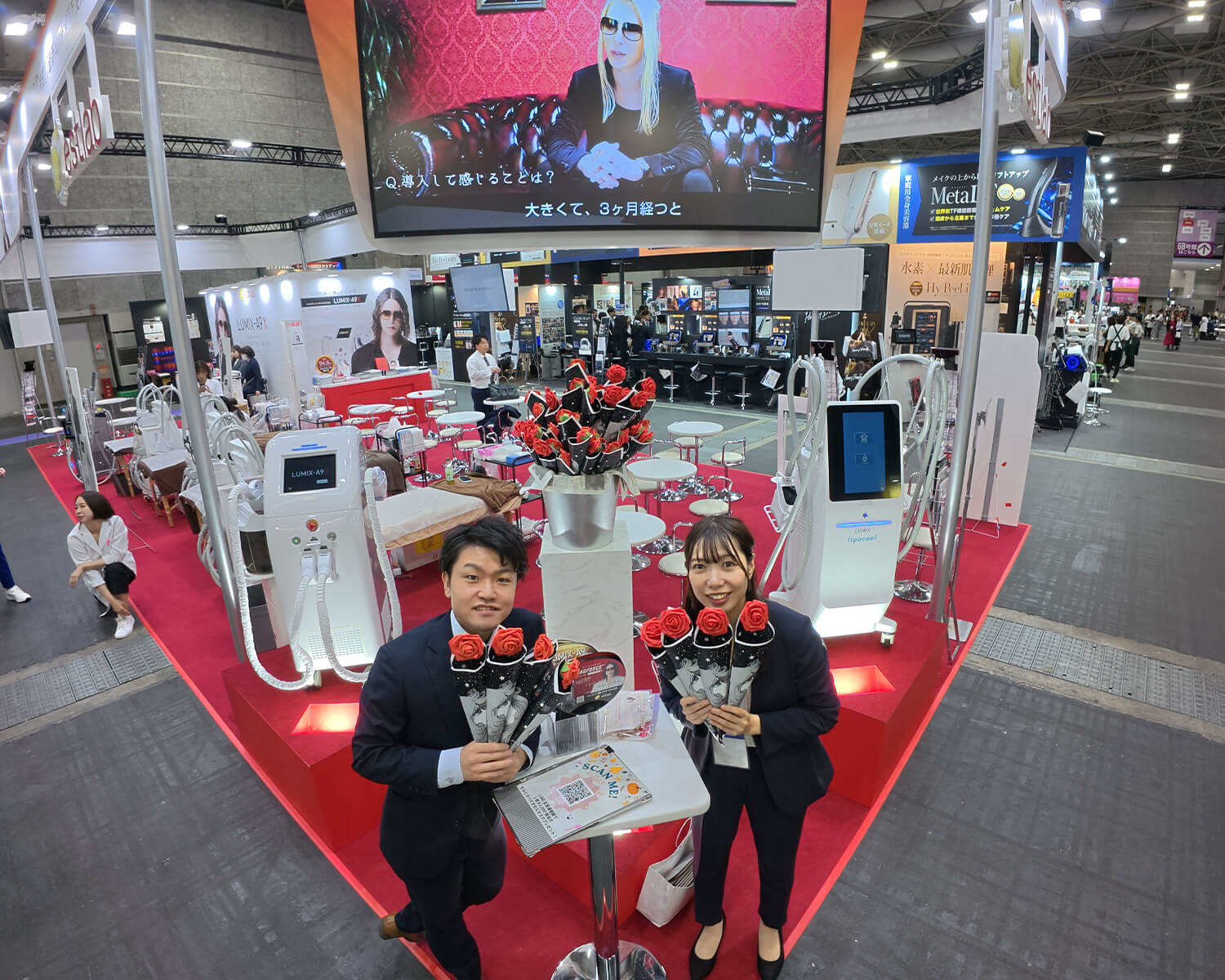 大規模な美容機器の展示会ブース「BEAUTY WORLD JAPAN」の全体像。赤いカーペットのブース中央に、バラの花束を持ったスーツ姿の男女スタッフ2名が立っています。背景には最新の美容機器や大型スクリーン、多くの来場者が行き交う会場の様子が見えます。