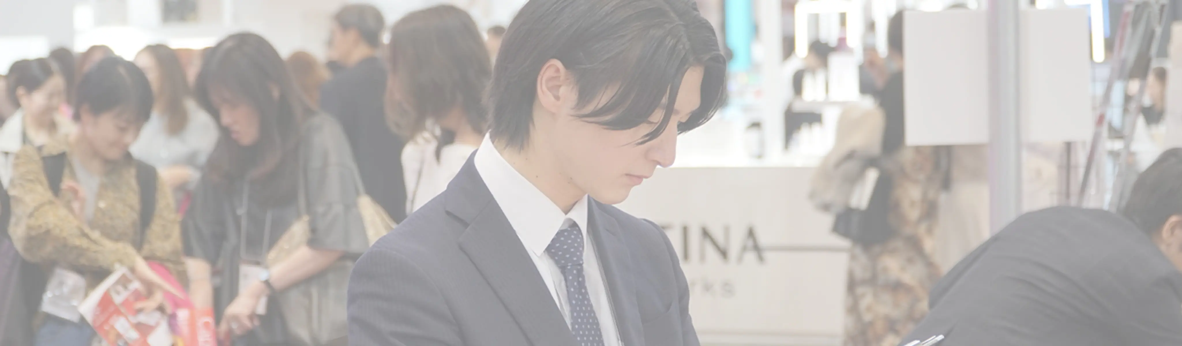 展示会で下を向いてメモを取るスーツ姿の男性と、背景にいる多くの来場者