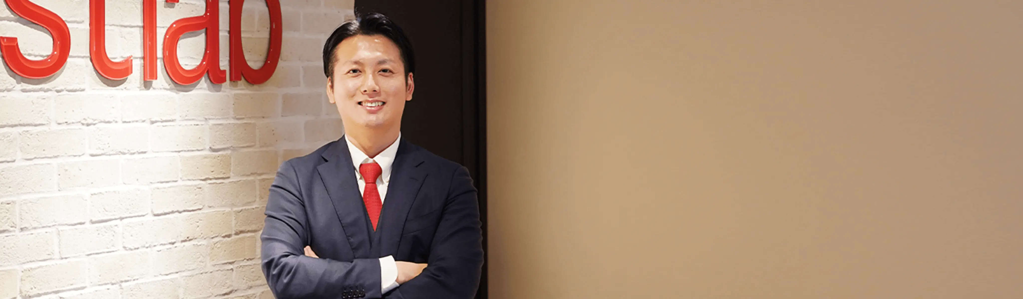「株式会社エストラボ」のオフィスで、ロゴを背に腕を組んで微笑む 代表取締役社長 木原 武士。赤タイがアクセントのビジネススタイル。