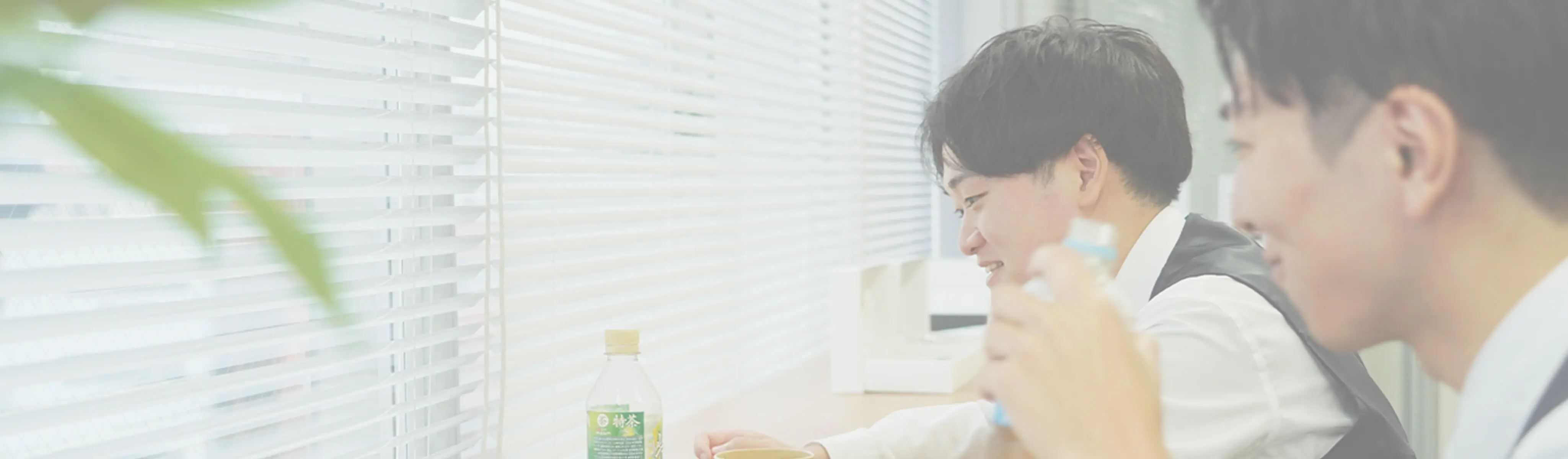 ブラインドの横のテーブルで、笑顔でペットボトル飲料を飲む男性とその同僚